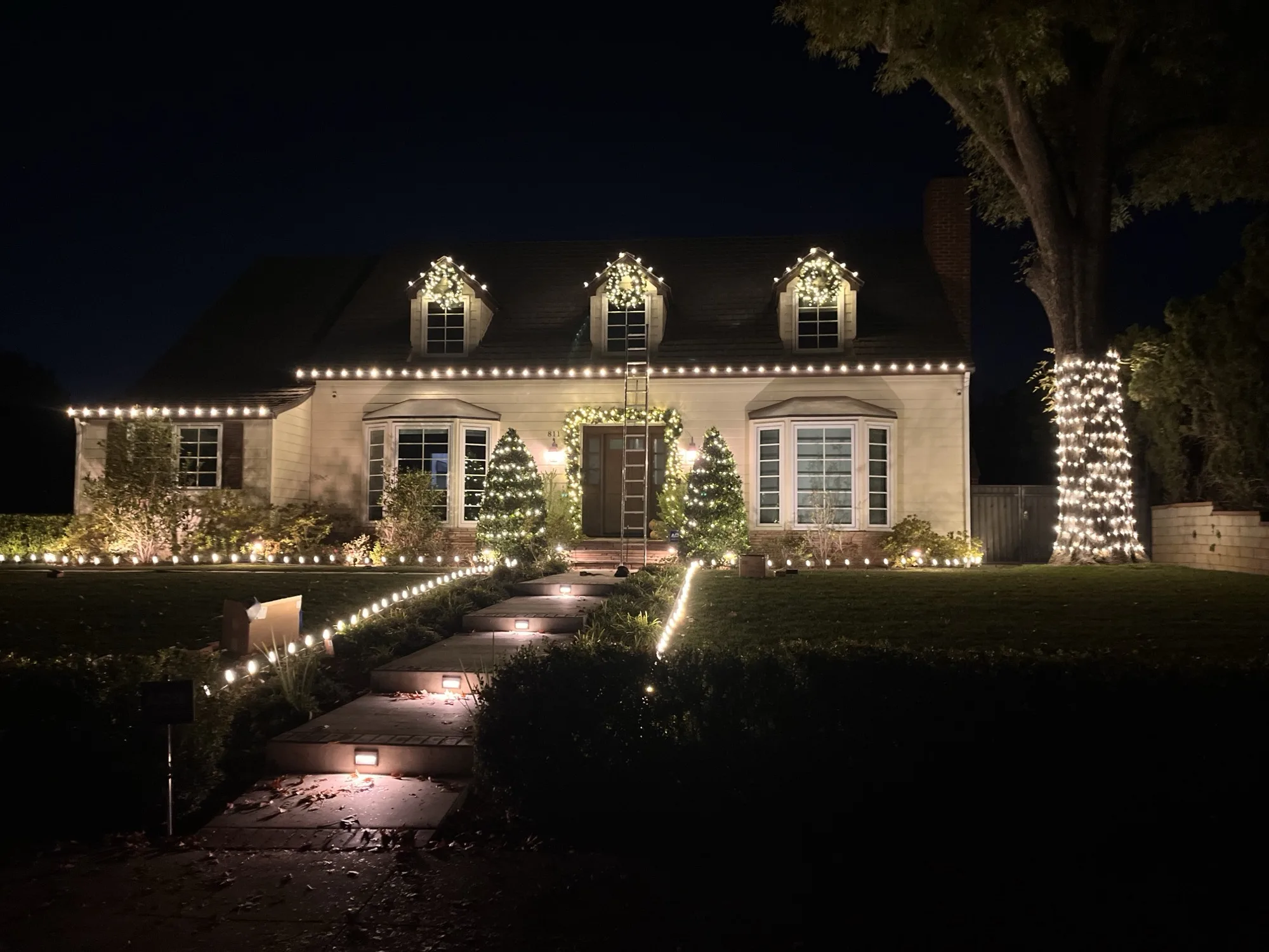 Cape Cod home with warm-white roofline lights and dormer wreaths by Bee Merry
