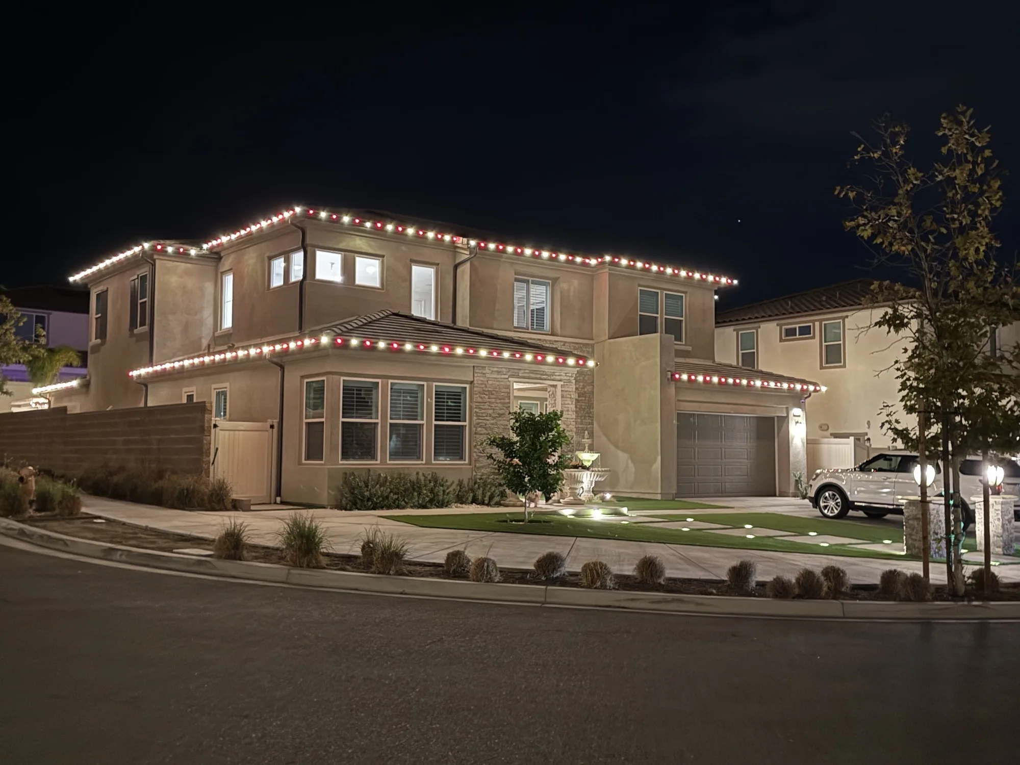 Stucco home with red and warm-white Christmas lights and a lit fountain by Bee Merry